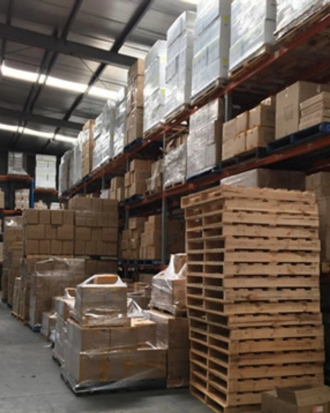 Warehouse interior with tall storage racks holding boxes, pallets, and shrink-wrapped goods under fluorescent lighting